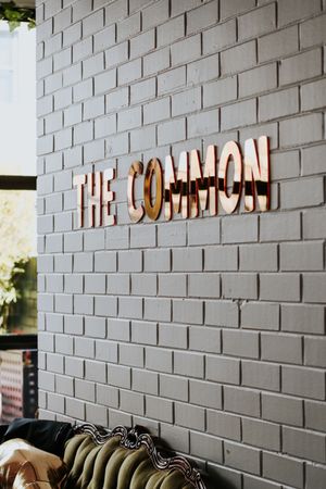 Gold letter signage reading The Common mounted on a gray painted brick wall above a vintage sofa.