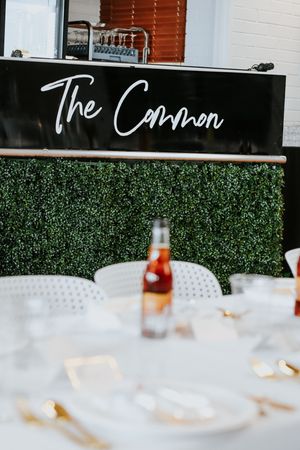 Elegant table setting at The Common venue with white chairs, gold cutlery, and a green hedge bar backdrop.