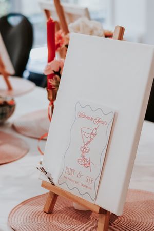 A Paint and Sip welcome sign on a wooden easel with a cocktail illustration at an elegant party setup.