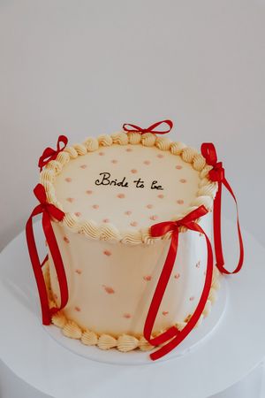 Elegant bride-to-be cake with cream frosting, red ribbon bows, and pink heart details on a white pedestal.