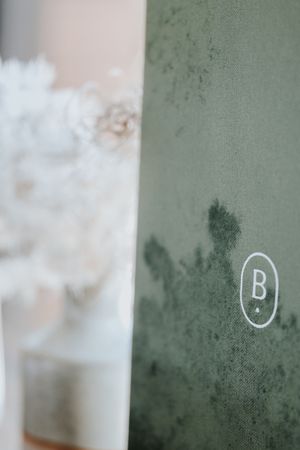 Close-up of a green linen book cover with a monogram letter B logo, beside soft white dried flowers.