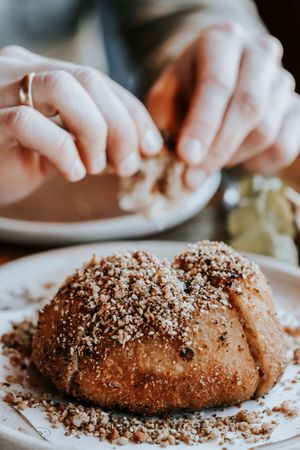 Hands sprinkling crushed nuts over a golden bread roll on a white plate.