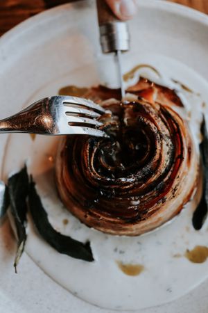 Close-up of a roasted onion on a white plate, being pierced by a fork with sage leaves and balsamic glaze.