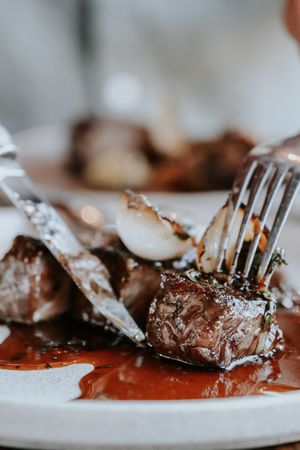 Close-up of juicy grilled steak pieces in rich red sauce on a white plate, served with fork and knife.