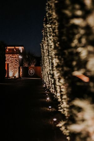 Nighttime venue entrance with warm string lights on hedges and illuminated stone wall with letter B sign.