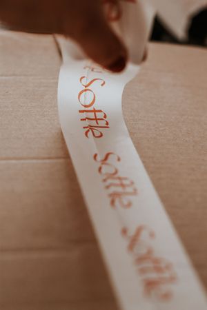 White satin ribbon with repeating copper script text Soffle being applied to a cardboard box.