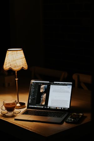 Cozy dark workspace with glowing lamp, laptop, coffee cup, and smartphone on a wooden desk at night.