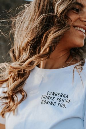 Smiling woman with wavy hair wearing a white tee reading Canberra Thinks You're Boring Too.