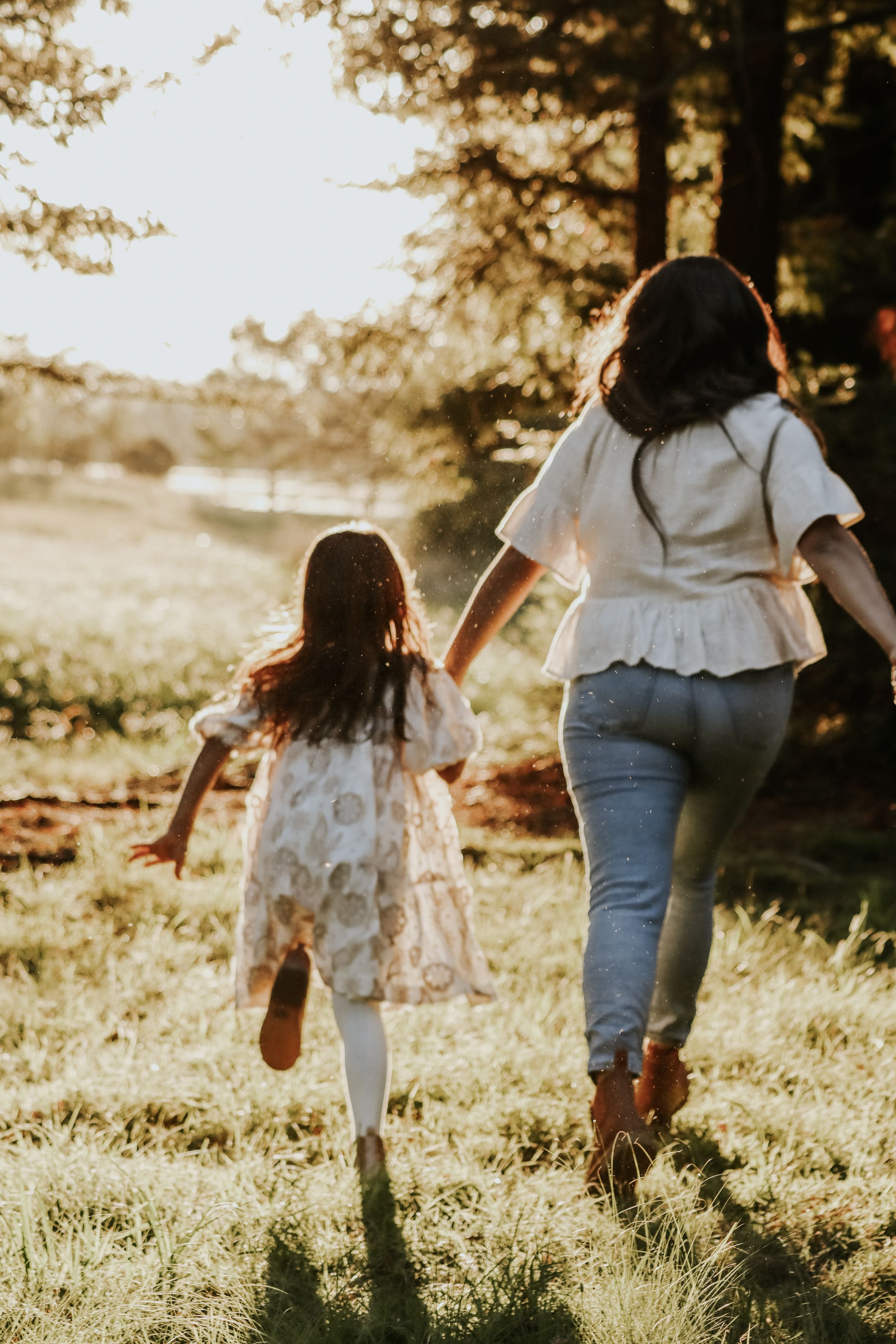 A mother and young daughter hold hands running through a sunlit forest meadow, both wearing white outfits.
