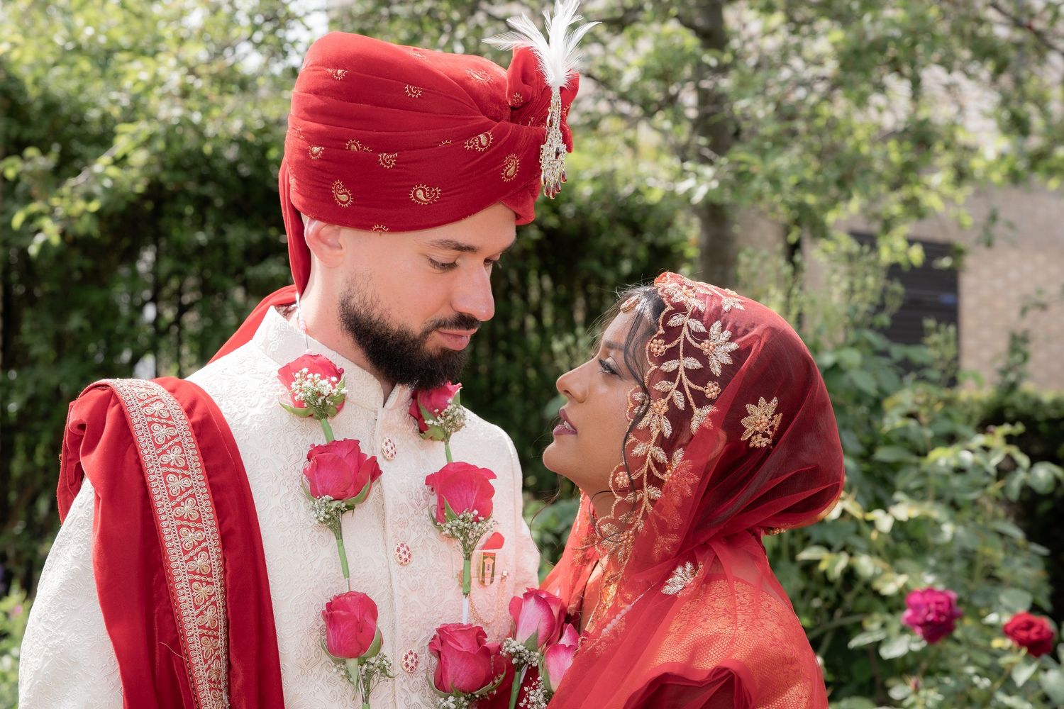 South Asian couple in traditional wedding attire, groom in white sherwani and red turban, bride in red dupatta, holding roses.