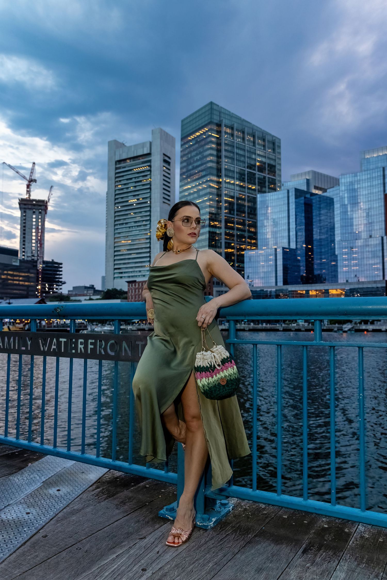 Woman in olive green dress poses on waterfront boardwalk with city skyline at dusk.