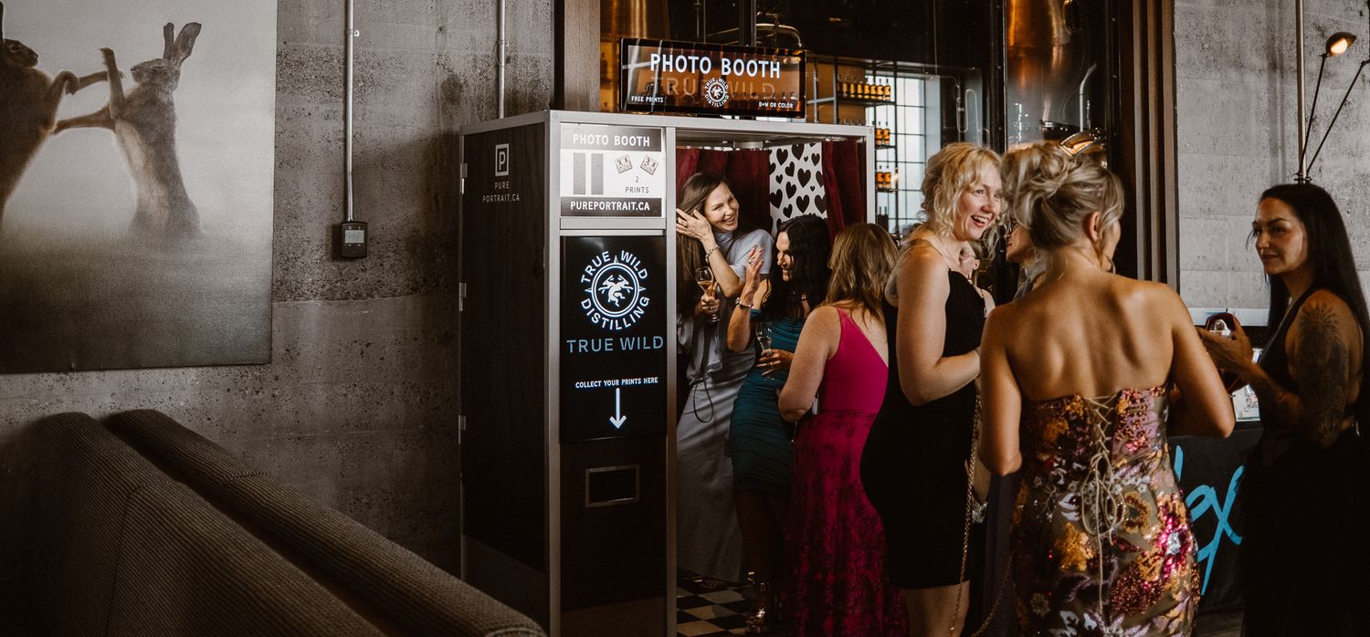 Guests gather near a photo booth and bar area at a dimly lit event venue with exposed brick walls.