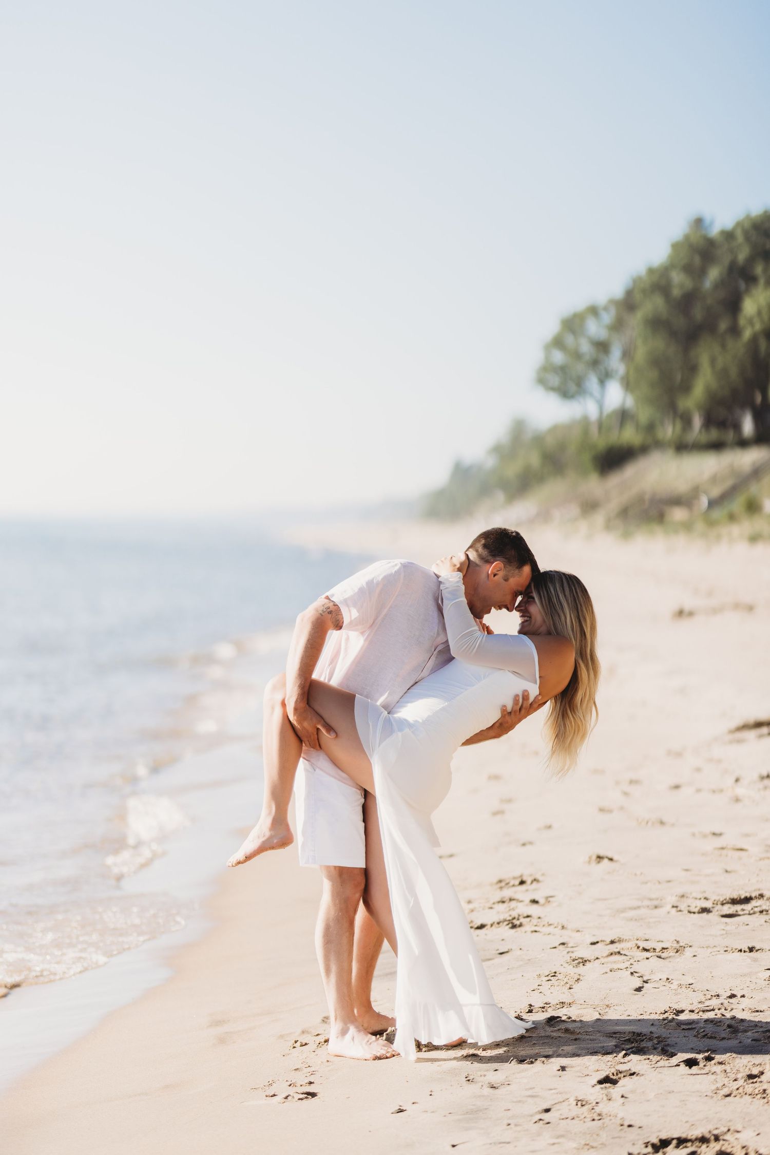 Couple in white outfits sharing a romantic kiss on a sunny sandy beach, man lifting woman near ocean shoreline.