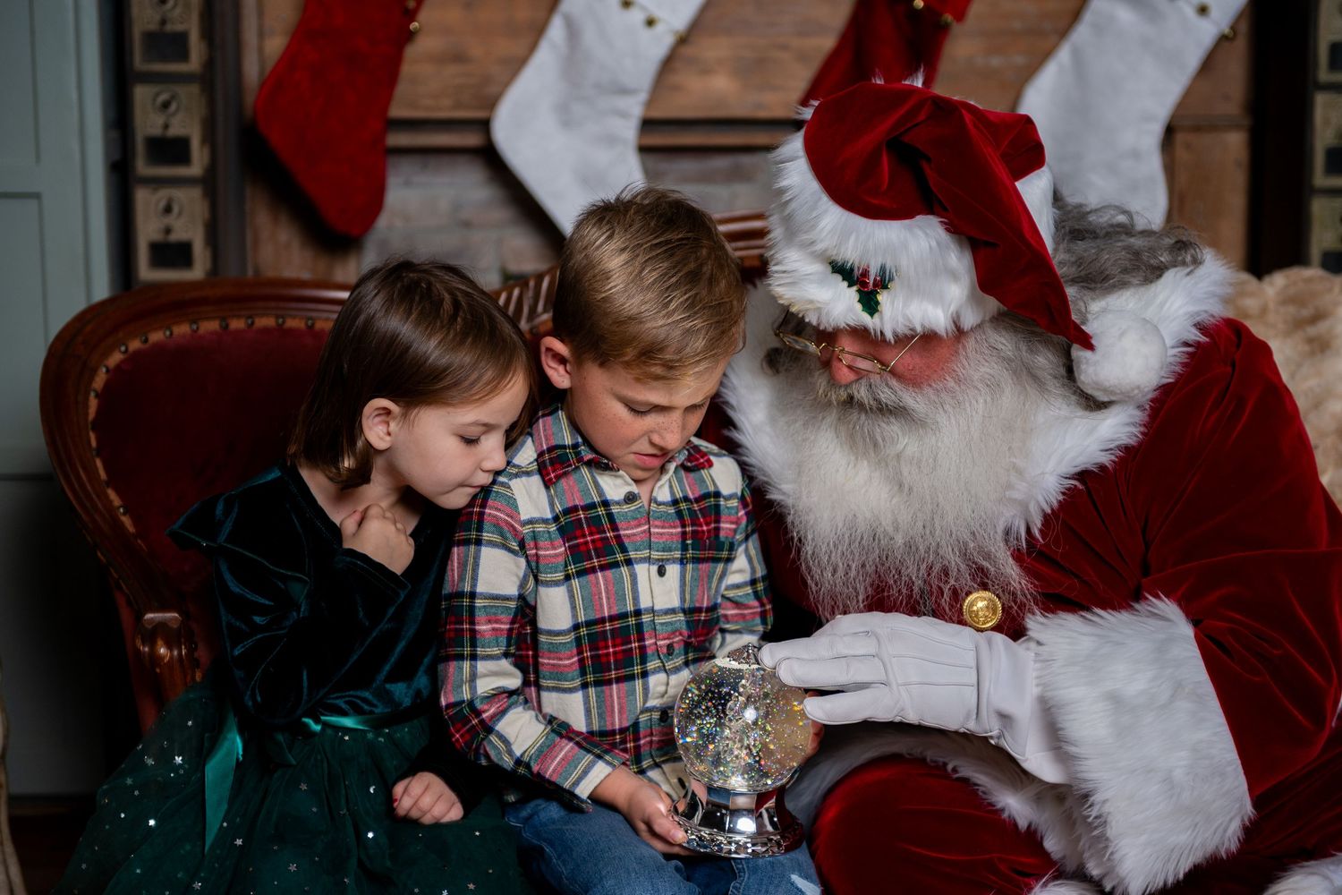 Two children sit with Santa Claus examining a snow globe during a festive Christmas photo session.