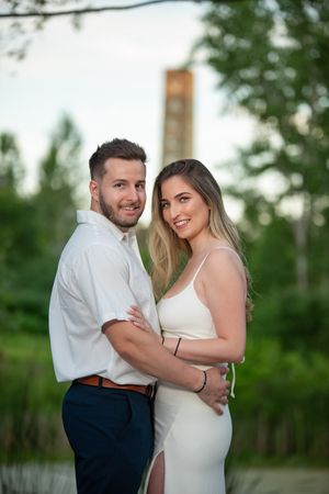 A smiling couple in white outfits embrace outdoors with lush green trees and a clock tower in the background.