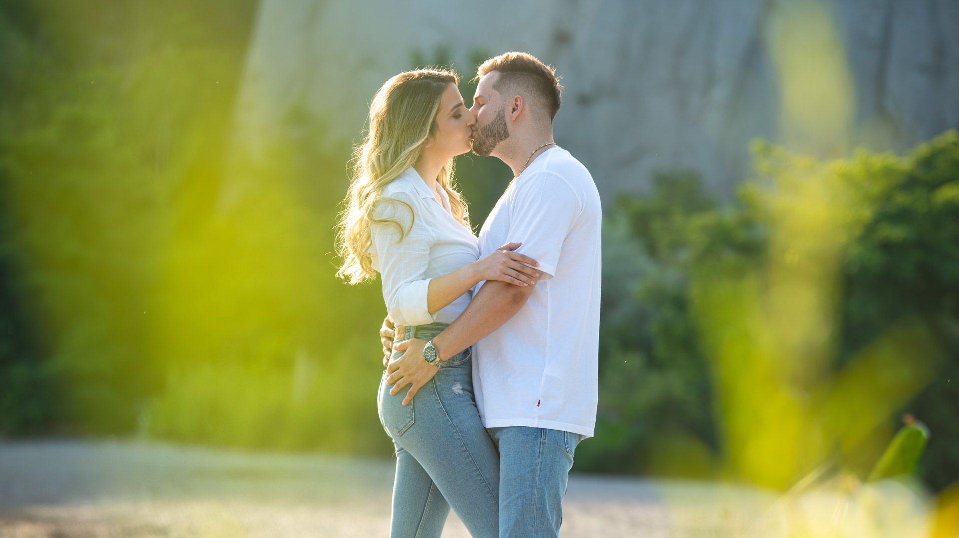 A couple shares a romantic kiss outdoors in warm golden sunlight with greenery in the background.