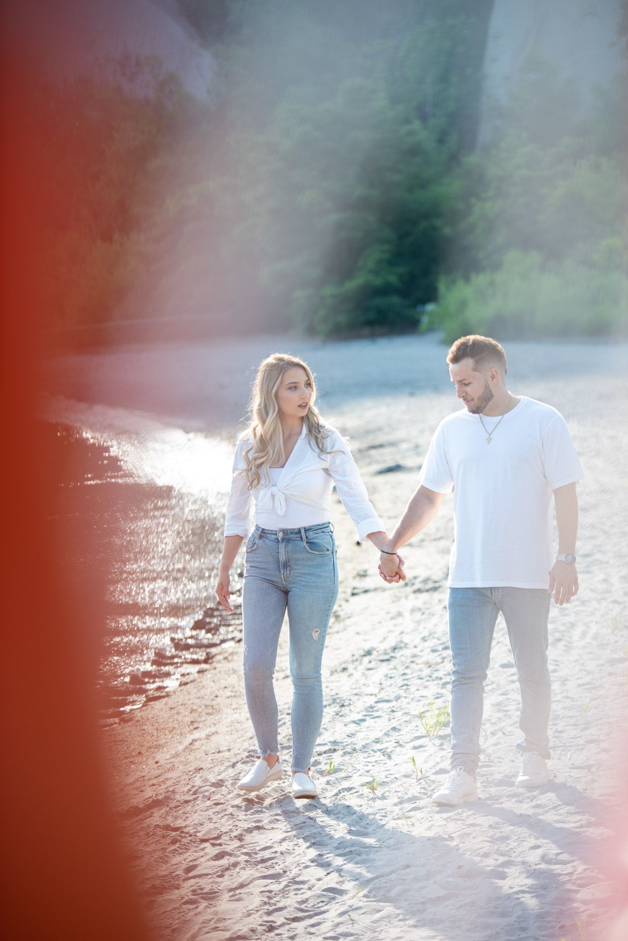 A couple holds hands while walking along a sunlit sandy path surrounded by green trees.
