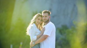 Happy couple embracing and smiling together outdoors with soft green bokeh and sunlight behind them.