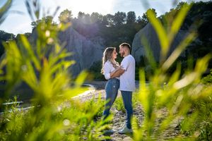 Couple facing each other tenderly near a rocky shoreline framed by vibrant green plants in sunlight.