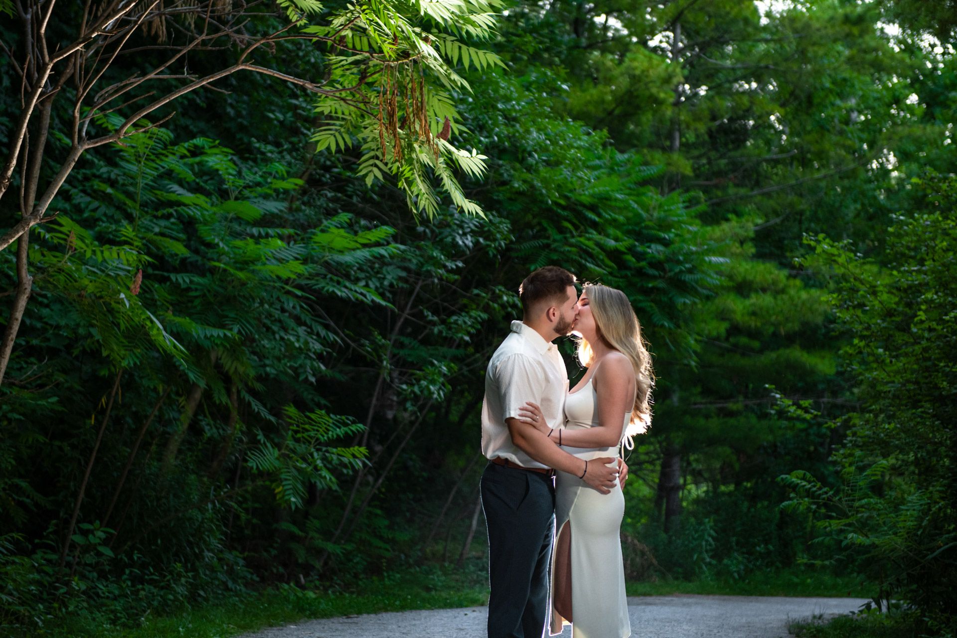 A couple shares a tender kiss surrounded by lush green trees during their outdoor engagement session.
