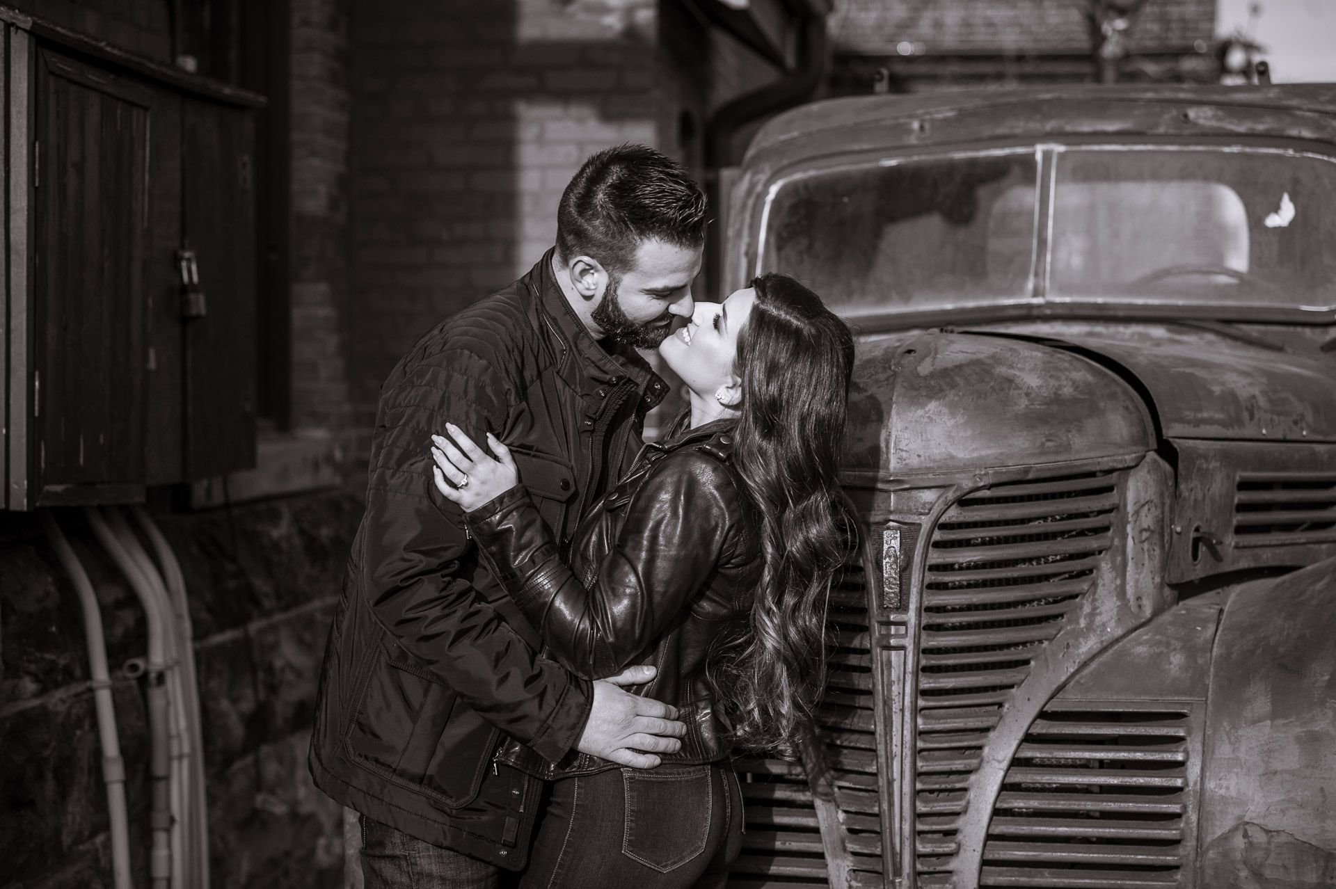 Black and white photo of a couple kissing beside a vintage rusty truck in a moody outdoor setting.