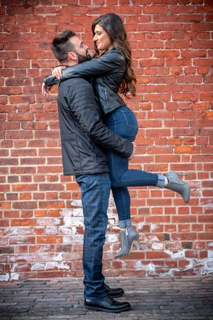 Couple embracing against a red brick wall, man lifting woman in matching black leather jackets and jeans.