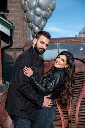 Couple posing together by a vintage rusty truck with silver balloons, both wearing black leather jackets.