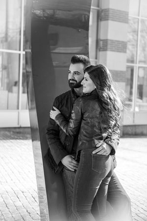 Black and white photo of a couple in leather jackets embracing near a reflective surface outdoors.