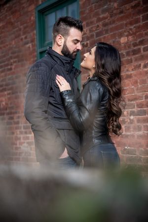 Couple in leather jackets gazing into each other's eyes near a rustic brick wall.