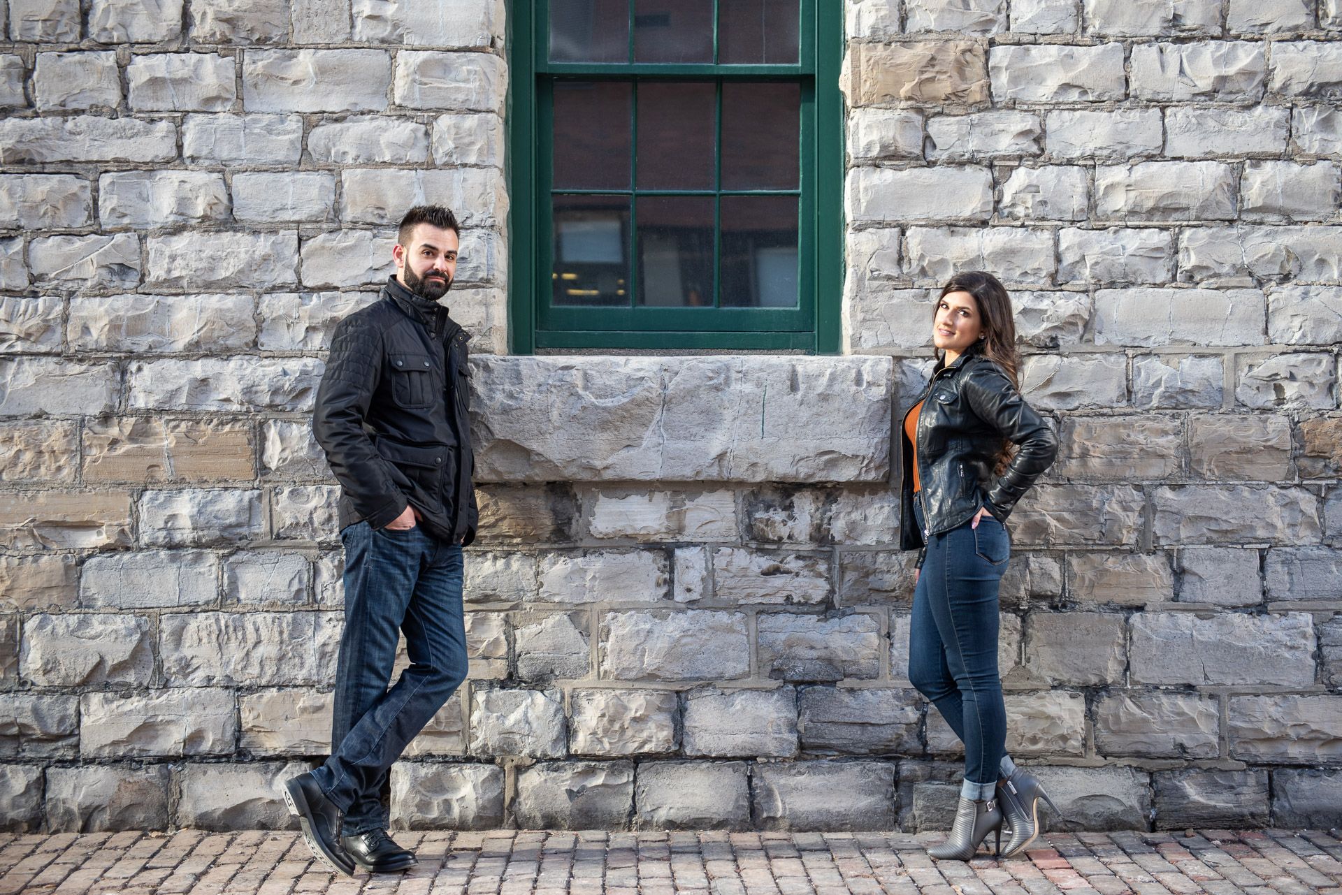 Man and woman posing casually apart against a historic stone wall with green window.