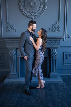 Stylish couple in formal wear sharing an intimate moment near an ornate grey fireplace.