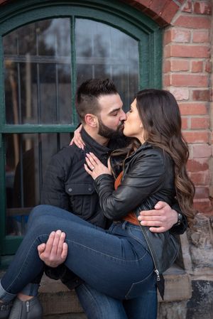 Couple sharing a tender kiss, man holding woman in his arms outside a brick building with arched window.