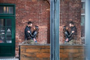 Couple sharing a tender moment reflected in glass beside a brick building with an Open sign.
