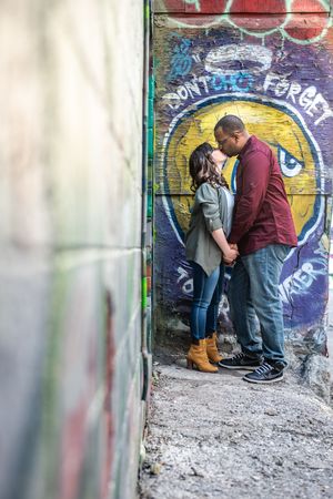 Couple sharing an intimate moment in a narrow alley with a yellow face graffiti mural behind them in Graffity Alley in Toronto.