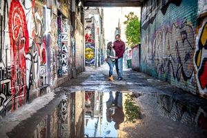 A couple embraces in a graffiti-lined alley with their reflection visible in a large rain puddle below in Graffity Alley in Toronto..