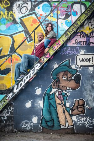 Couple sitting on graffiti-covered stairs next to a cartoon dog mural saying WOOF in an urban alley in Graffity Alley in Toronto..