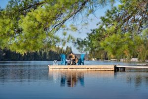 Couple sitting on a lake dock with blue Adirondack chairs surrounded by lush green pine trees.
