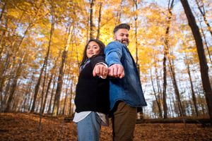 Couple standing back-to-back in autumn forest holding hands with golden foliage behind them.