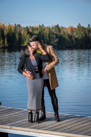 Couple sharing a kiss on a wooden dock with a blue lake and fall trees behind.