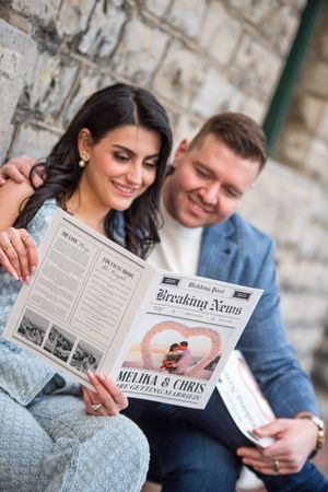 Engaged couple smiling while reading a Breaking News newspaper announcing Melika and Chris are getting married.