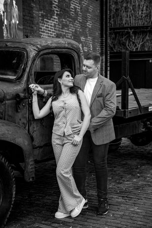 Black and white photo of a couple holding hands and smiling beside a vintage rusty truck.