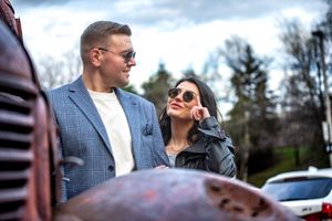 Stylish couple wearing sunglasses standing close together near a vintage rusty vehicle outdoors.