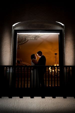 Bride and groom silhouetted in a glowing arched doorway on a balcony at night.