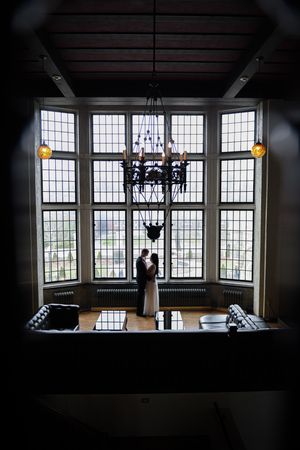 Wedding couple embracing before grand leaded glass windows with chandelier above.
