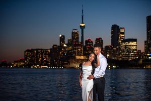 A couple embraces romantically at night with the illuminated Toronto city skyline reflecting on the water.