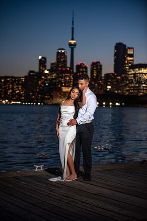 Couple embracing on a waterfront dock at night with the illuminated Toronto city skyline reflected on the water.