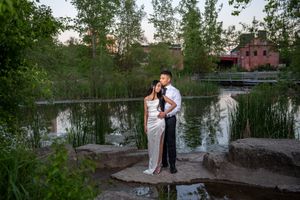 A couple stands close together on rocks beside a serene pond with greenery and buildings behind them.