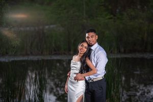 A couple embraces warmly beside a calm lake at dusk surrounded by tall reeds and green trees.