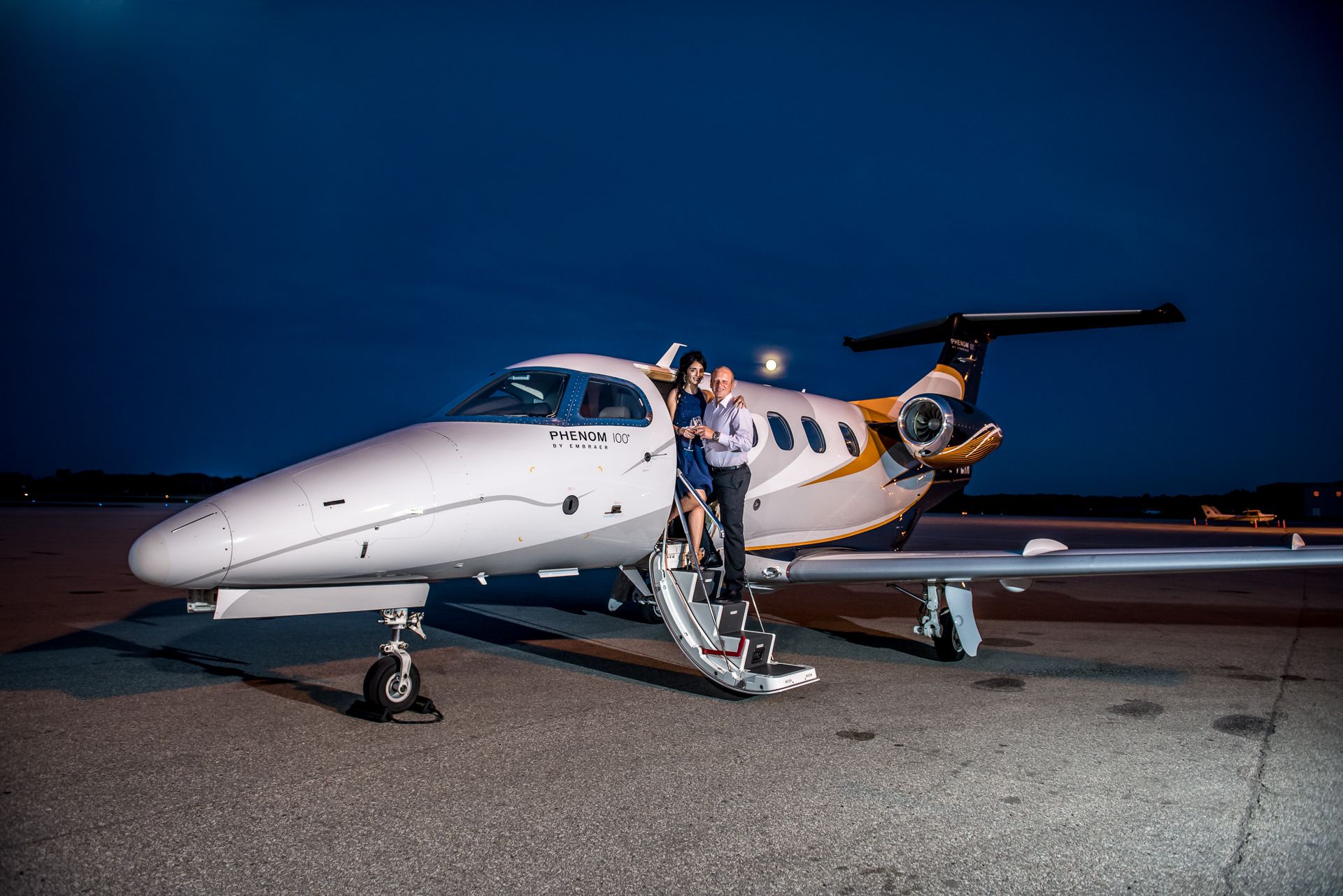 Couple posing together on stairs of a white private jet on a tarmac at dusk.