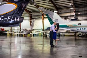 Couple embracing romantically in an aircraft hangar surrounded by private jets.